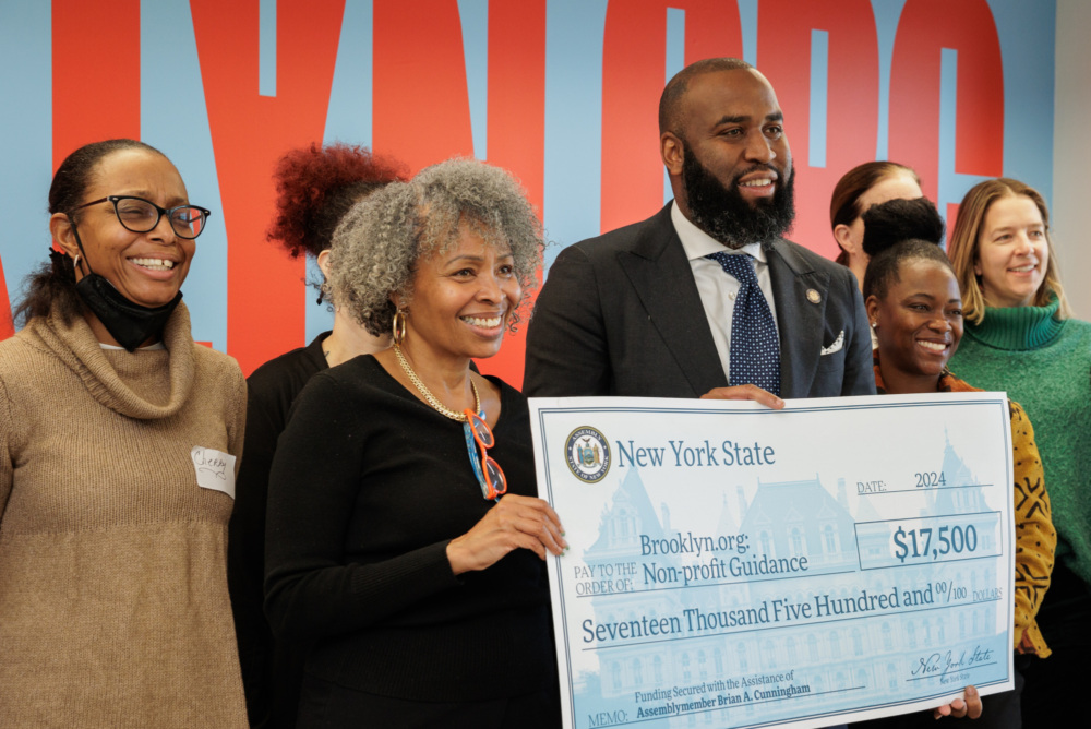 A diverse group of people smiling and holding a large ceremonial check for $17,500 from New York State, with a colorful wall in the background.