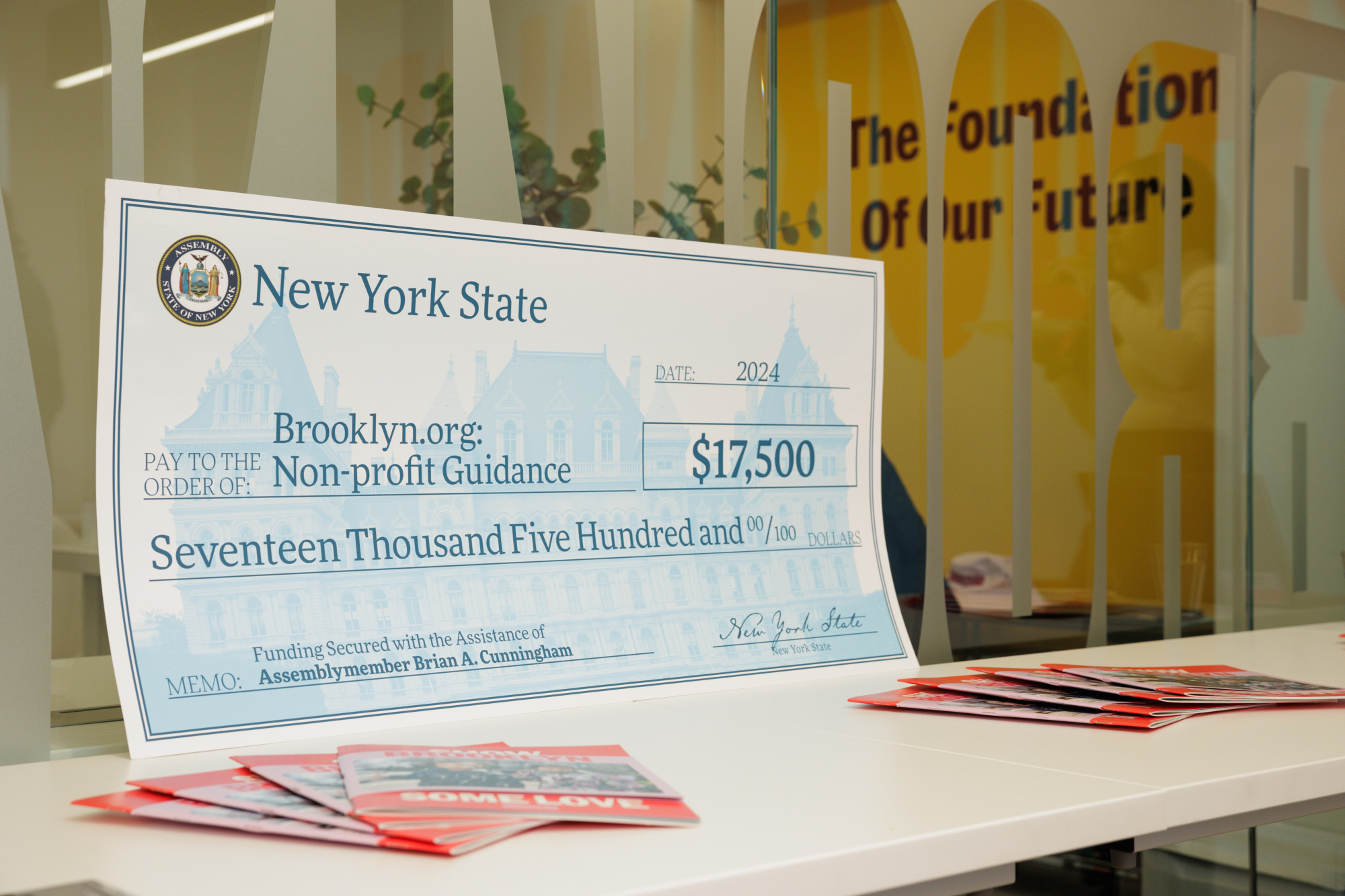 A large ceremonial check for $17,500 from New York State to Brooklyn.org for non-profit guidance, displayed on a table with brochures.