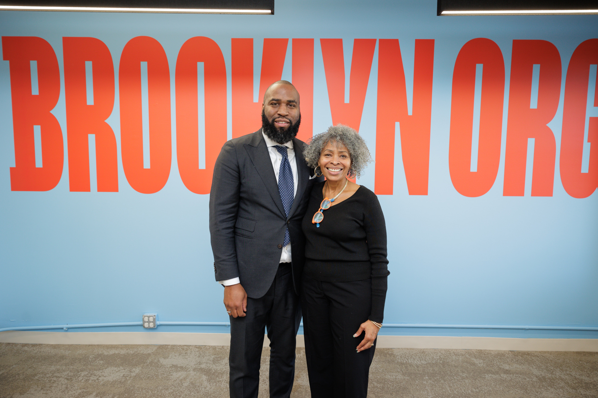 Two people standing and smiling in front of a wall with large red text that reads "BROOKLYN ORG.