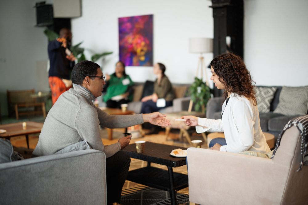 Two people sitting across from each other in a lounge, shaking hands. Other people are in the background on couches, and there's a colorful painting on the wall.