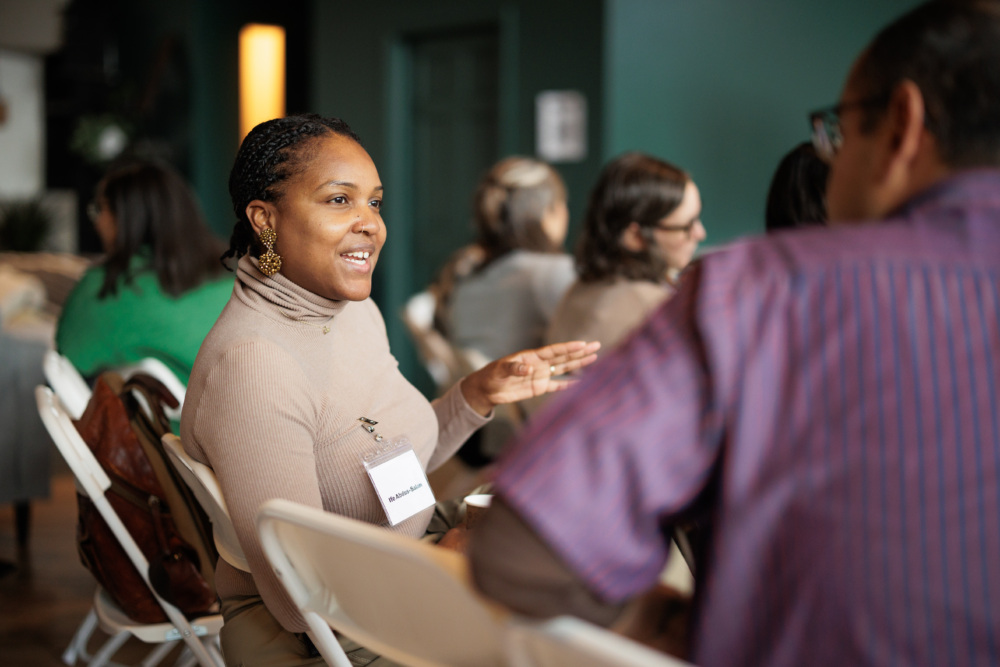 A woman wearing a name tag is engaged in conversation with a man in a purple shirt. They are seated in white chairs at an indoor event with other attendees in the background.