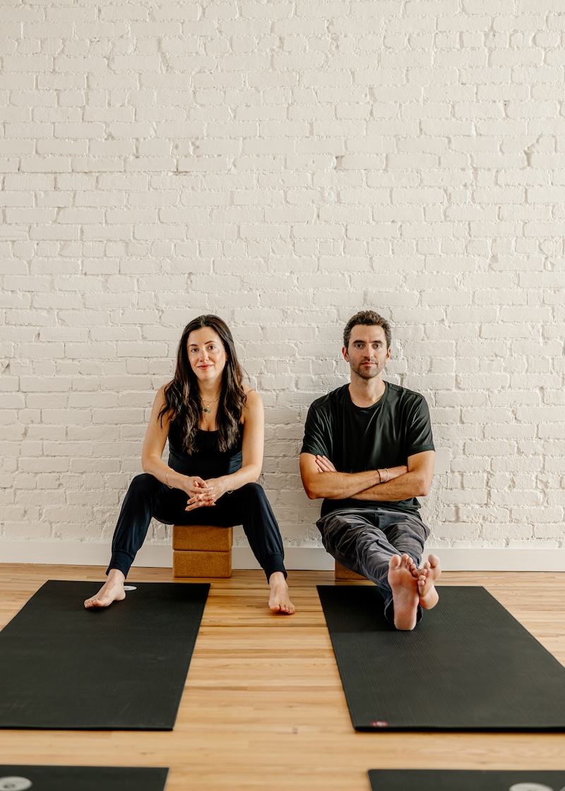 Two people sitting on yoga mats against a white brick wall. The person on the left sits on a block, while the person on the right sits cross-legged, both barefoot.