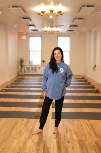 A woman with long brown hair stands barefoot in a yoga studio with wooden floors and yoga mats, wearing a blue sweatshirt and black pants, smiling at the camera.