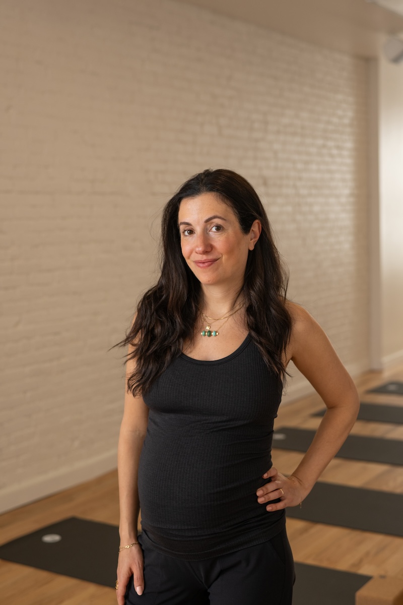 Person in a black tank top standing in a yoga studio with a hand on their hip. Yoga mats are visible on the wooden floor. White brick wall in the background.