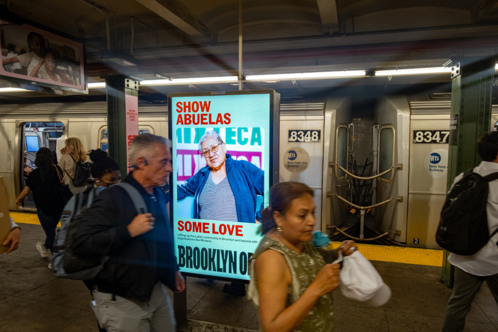 People walking on a subway platform with an advertisement reading "Show Abuelas Some Love" in the background.