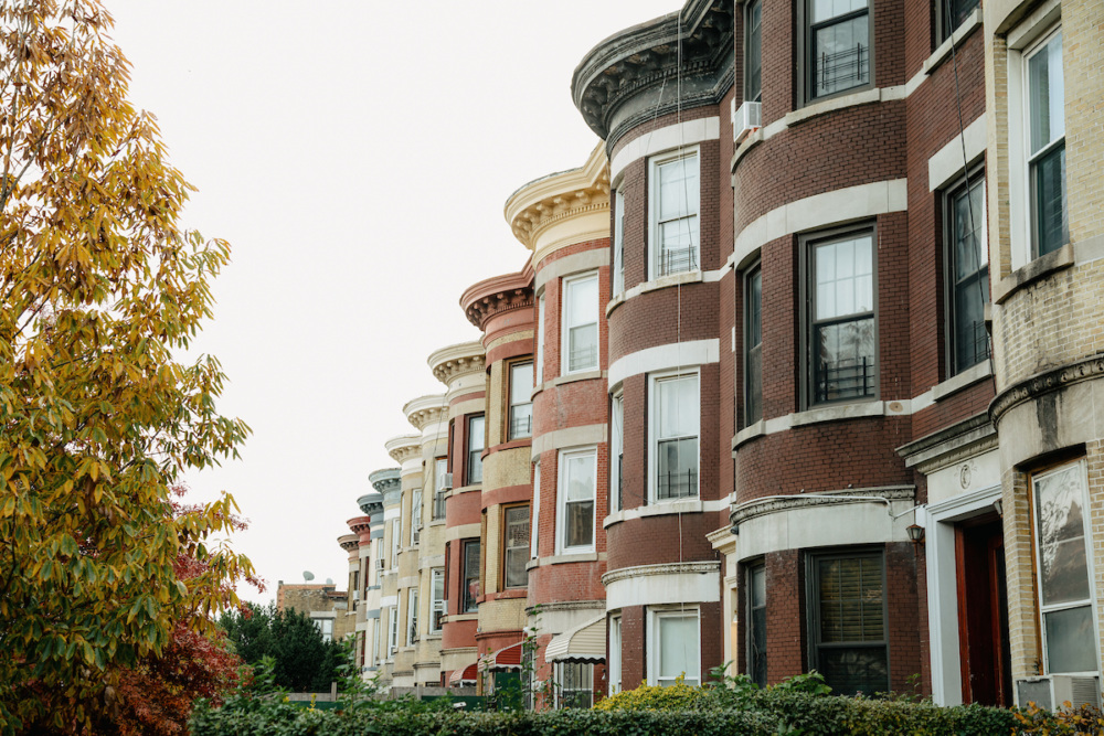 A row of historic brick row houses with rounded facades, featuring large windows and decorative details, alongside autumn-colored trees.