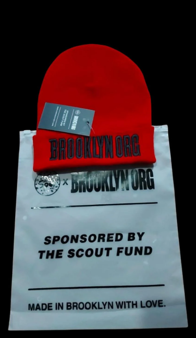 A red beanie with "Brooklyn.org" text placed on a white bag reading "Sponsored by the Scout Fund. Made in Brooklyn with Love." A tag is attached to the beanie.