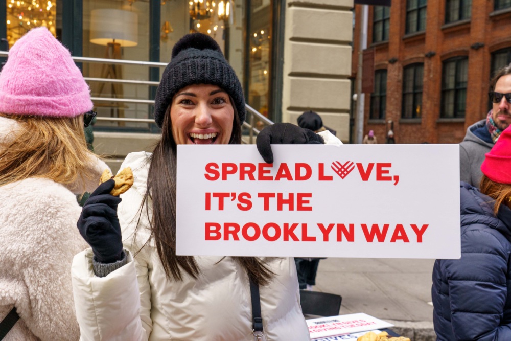 Person in winter clothing holds a sign reading "Spread Love, It's the Brooklyn Way" on a city street, smiling and holding a cookie.