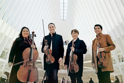 A quartet of musicians with string instruments stands in a bright, modern architectural space, smiling at the camera.