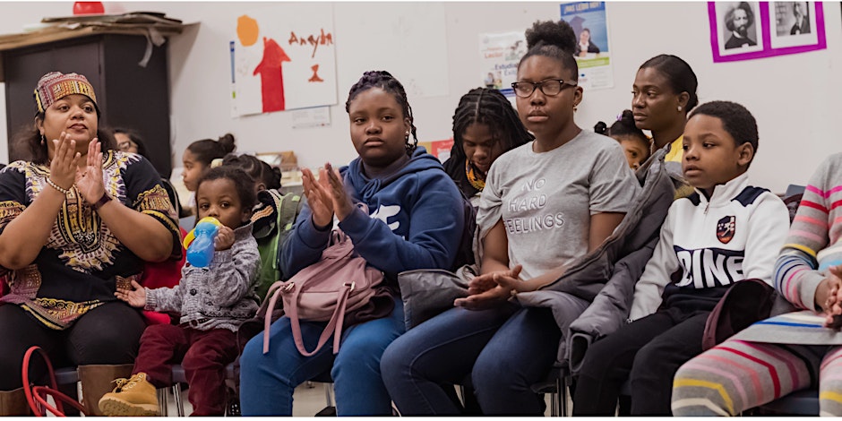 A group of people, including children and adults, sit in a classroom setting. Some are clapping, and a child is holding a sippy cup. Posters are visible on the walls.