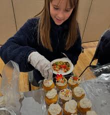 A young girl wearing a dark sweater and gloves is serving a plate of food, surrounded by cupcakes decorated with frosting and sprinkles.