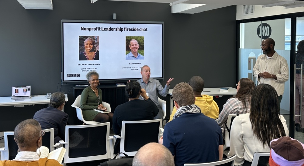 People attending a nonprofit leadership fireside chat in a modern conference room, with speakers seated and audience listening attentively.