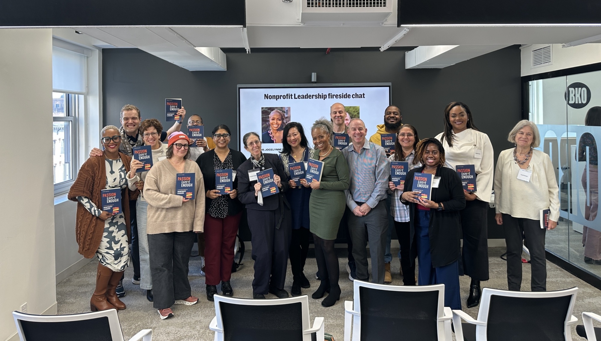 A group of people posing with books in front of a screen displaying "Nonprofit Leadership fireside chat" in a modern conference room.