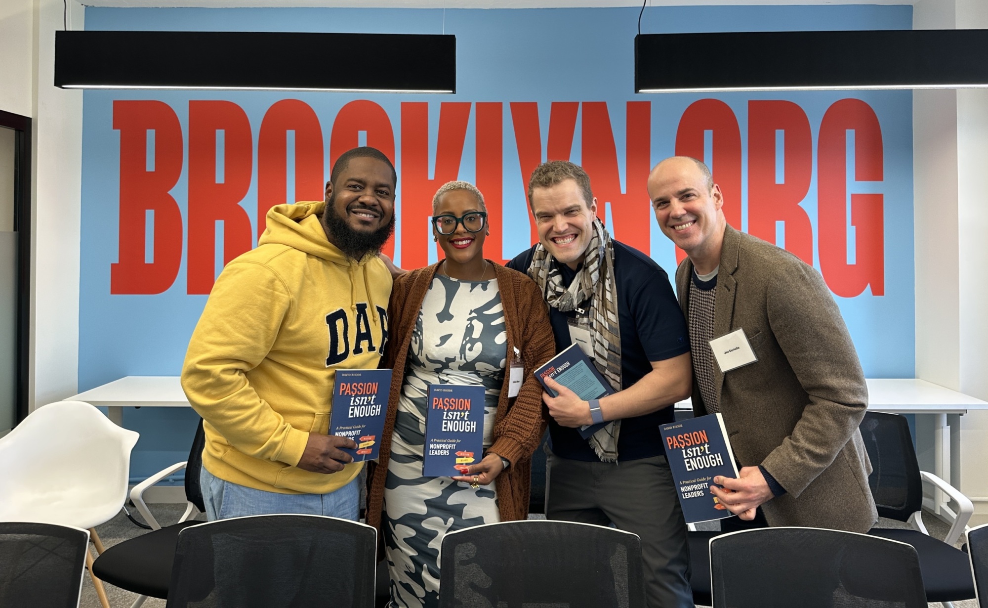 Four people in a room with a "BROOKLYN.ORG" sign in the background, holding books titled "Passion Isn’t Enough," and smiling at the camera.