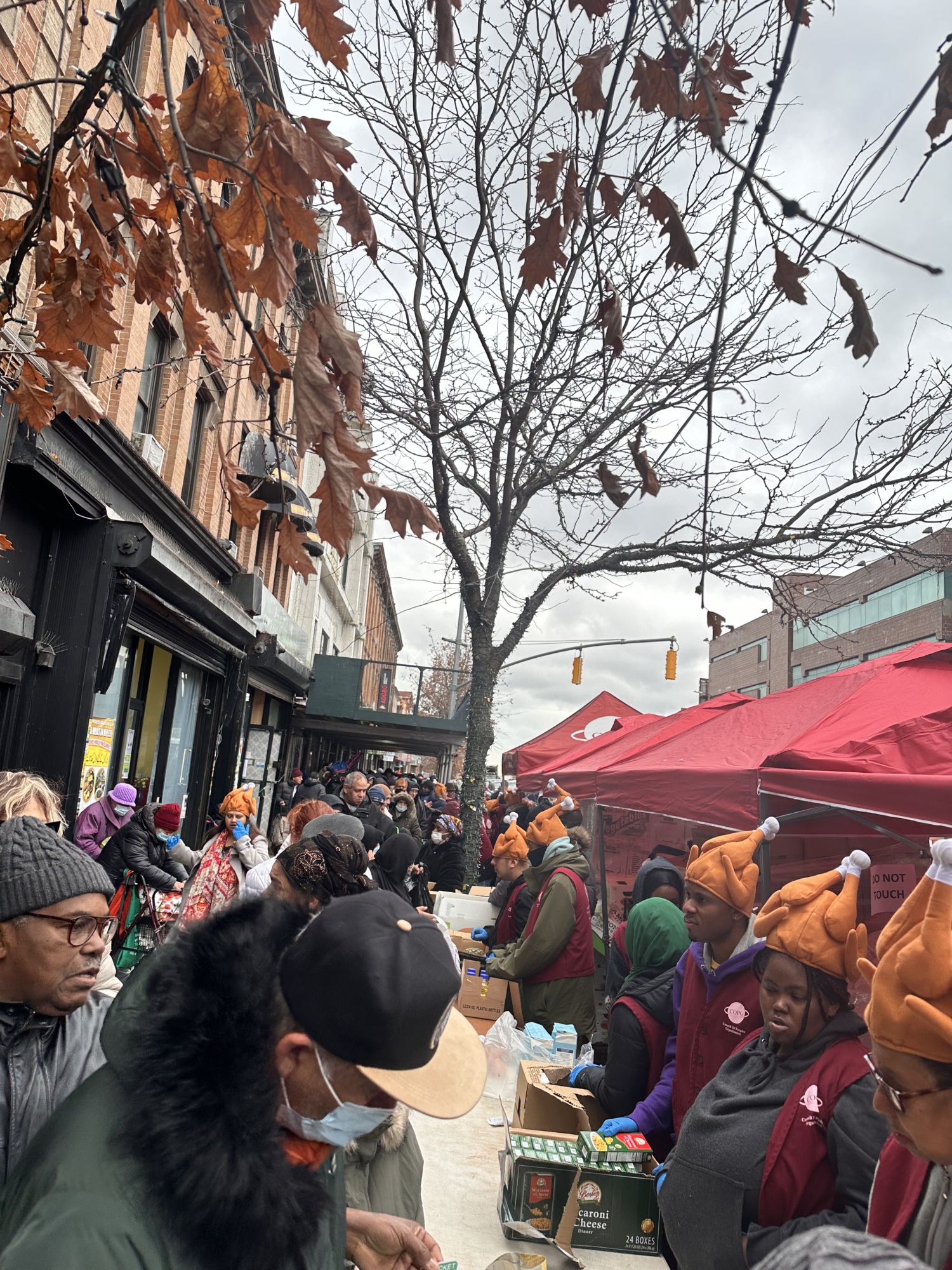 People gathered at an outdoor market with tables of goods, under trees with brown leaves. Some people wear hats shaped like turkeys.