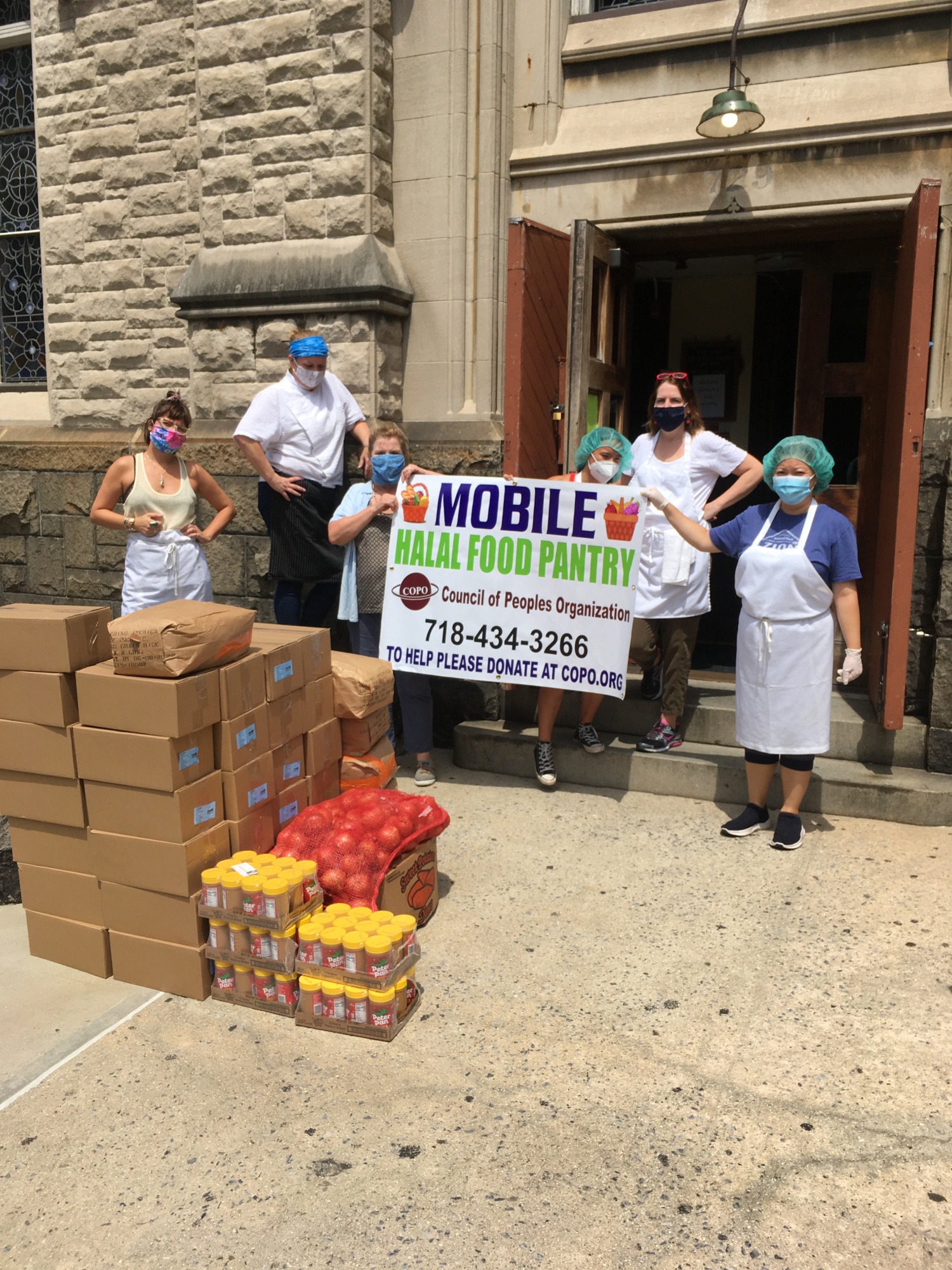 A group of people wearing masks and aprons stand outside a building holding a "Mobile Halal Food Pantry" banner. Boxes and bags of food are stacked nearby.