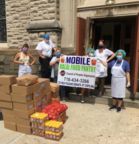 A group of people wearing masks and aprons stand outside a building holding a "Mobile Halal Food Pantry" banner. Boxes and bags of food are stacked nearby.