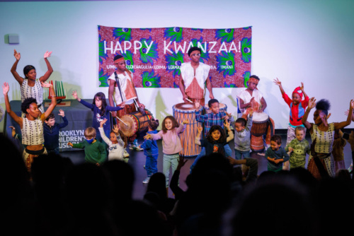 Performers and children celebrate Kwanzaa on stage with a colorful backdrop and drums.