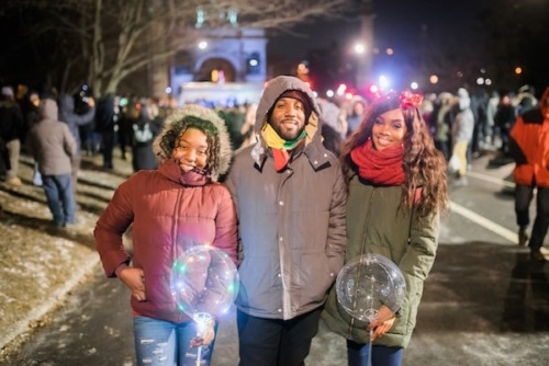 Three people in winter coats stand smiling at a nighttime outdoor event, holding clear balloons with lights inside.