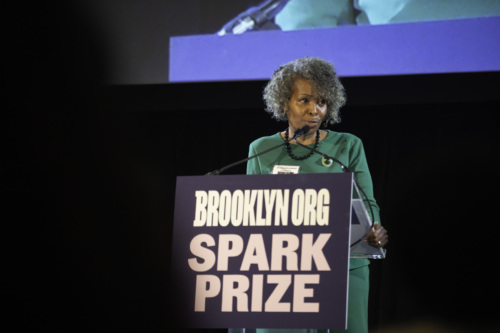 A woman in a green outfit speaks at a podium with a sign reading "Brooklyn.org Spark Prize.