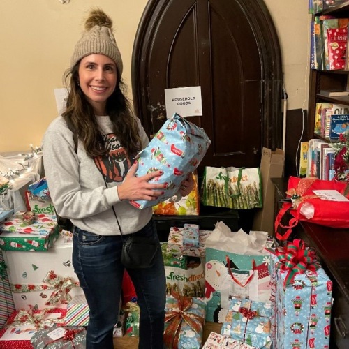 A person holding a gift stands in a room filled with wrapped presents and shopping bags.