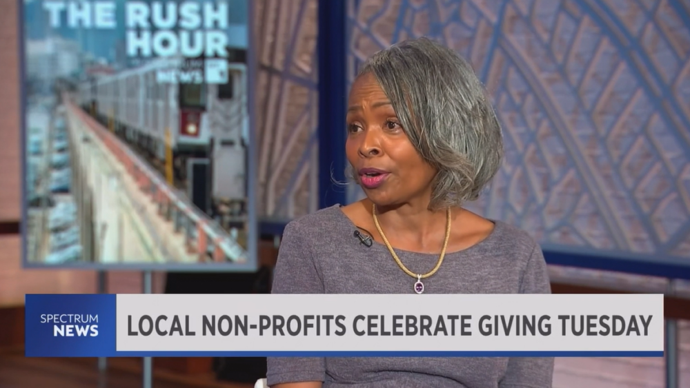 A woman with gray hair in a gray top speaks in a TV studio. A backdrop shows "The Rush Hour." The screen banner reads, "Local Non-Profits Celebrate Giving Tuesday.