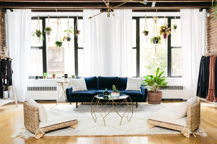 Modern living room with a blue sofa, hexagonal tables, plants hanging near large windows, and wicker chairs on a white rug.