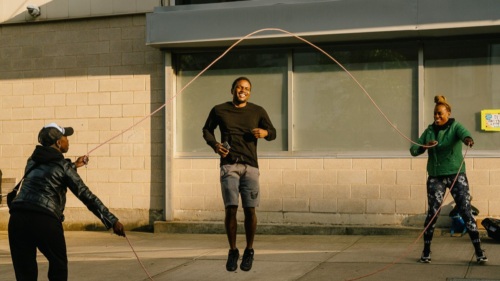 Three people are outdoors jumping rope: one man in the center jumping, and two others holding the rope ends on each side.