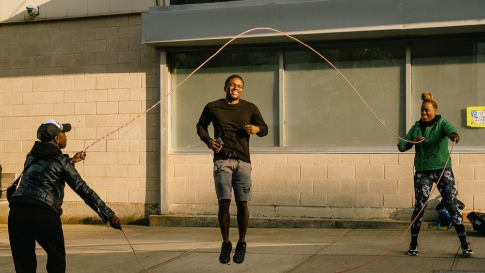 Three people playing jump rope outside a building, with one person jumping and two holding the rope.