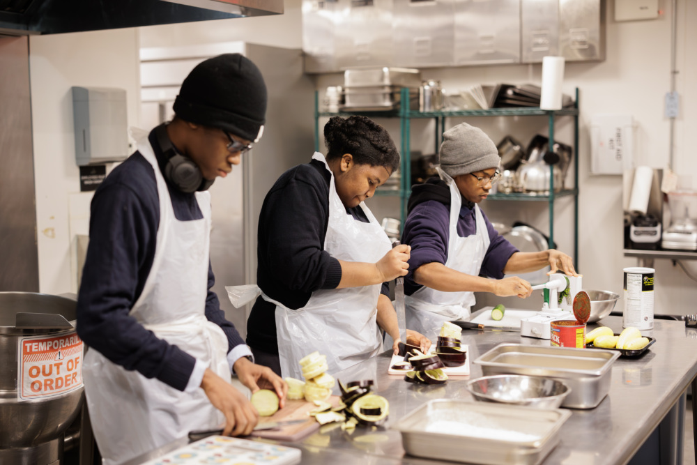 Three people in a kitchen wearing aprons and hats prepare food at a counter. One person is peeling, another is seasoning, and the third is opening a can. Cooking utensils and ingredients are visible.