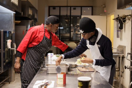 Two people in a kitchen preparing food. One wears a red chef coat and black striped apron, the other a dark sweater and white apron. Various cooking supplies are on the stainless steel counter.