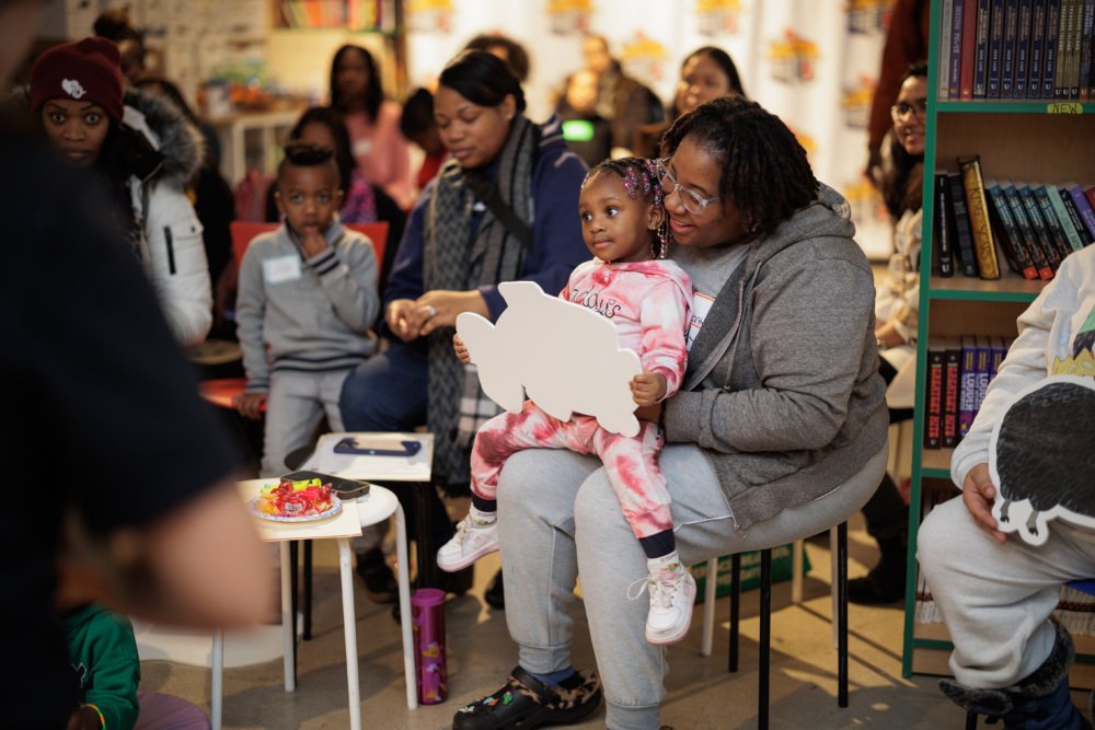 A gathering of people in a room, with a woman holding a child who has a cutout of an animal. Books are visible on shelves, and a small table with food is nearby.