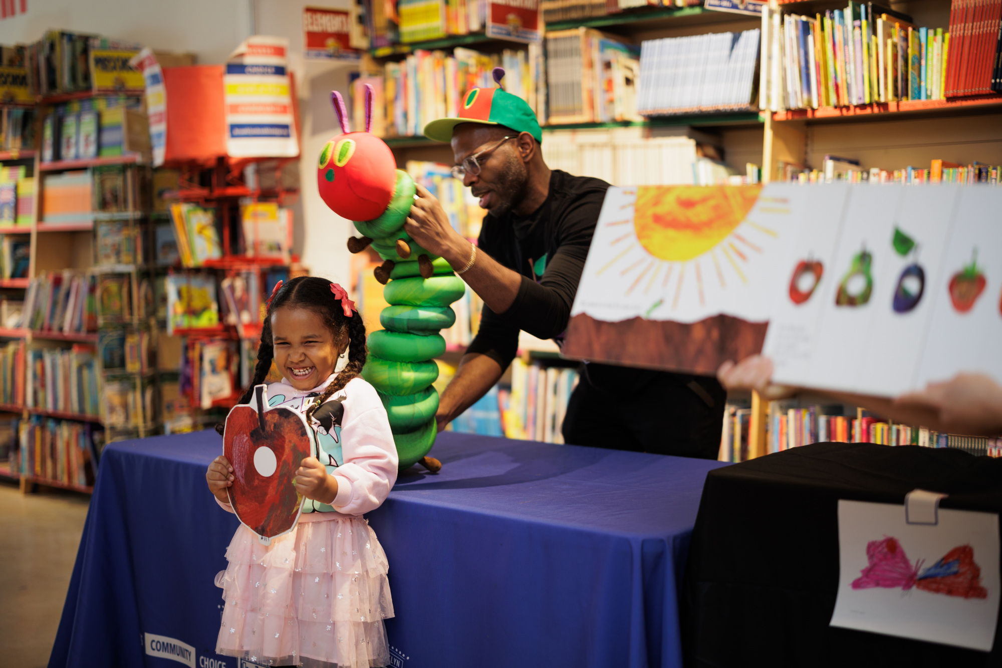 A child holds a caterpillar cutout while a person displays a large caterpillar puppet on a table. Bookshelves are in the background.