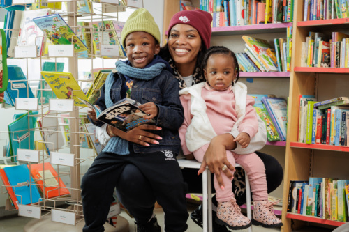 A woman and two children sit in a library. One child holds a comic book. Shelves filled with colorful books are in the background.