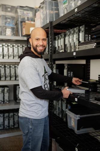 A man in a gray t-shirt and jeans smiling and organizing computer equipment on shelves in a storage room.