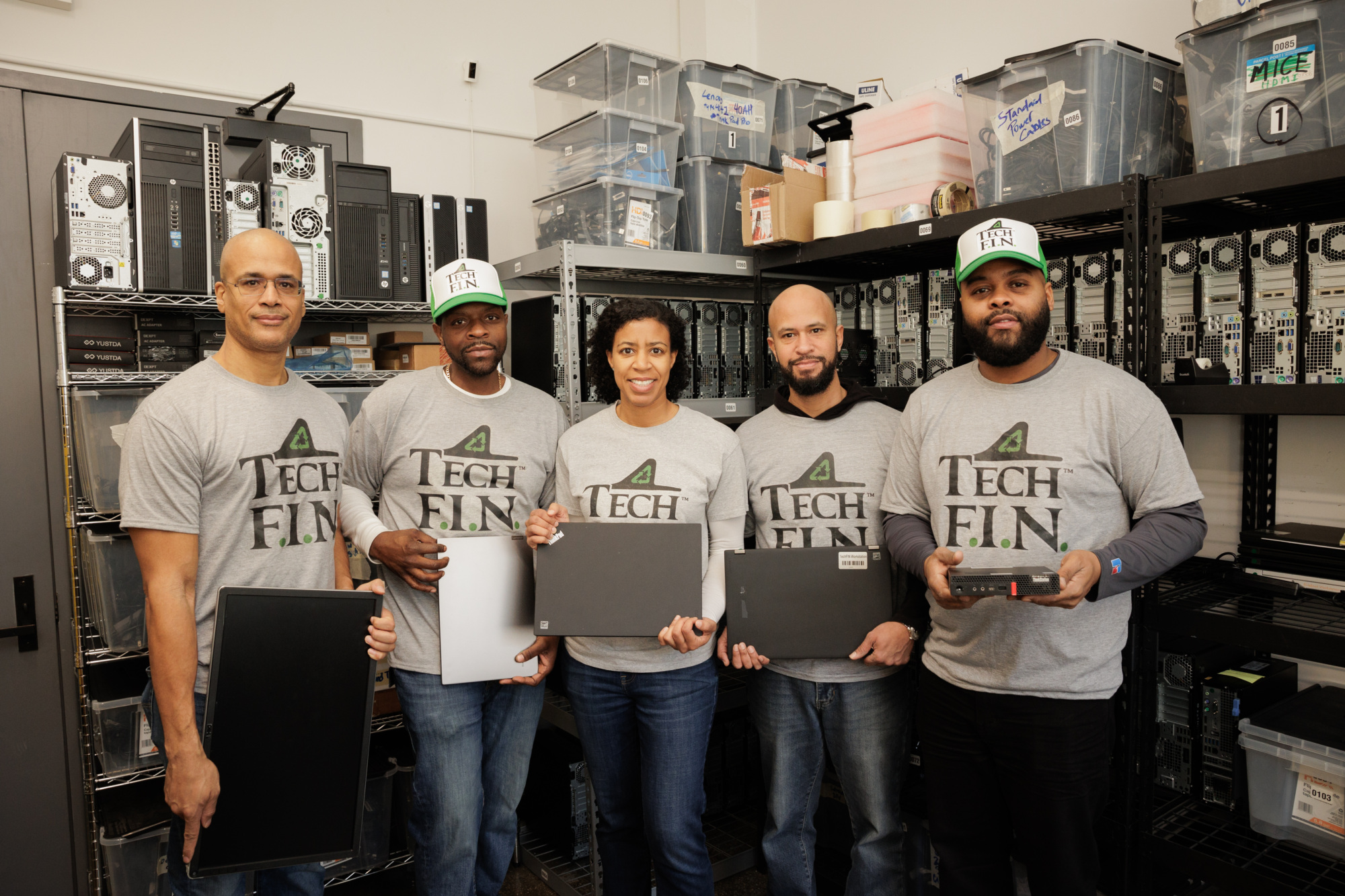 Five people in matching "Tech F.I.N." shirts stand in a room with shelves holding computer equipment, each holding a laptop or device.