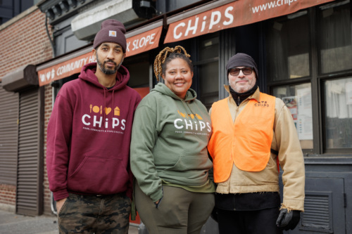 Three people stand together smiling outside a building with a "CHiPS" sign, wearing branded clothing.