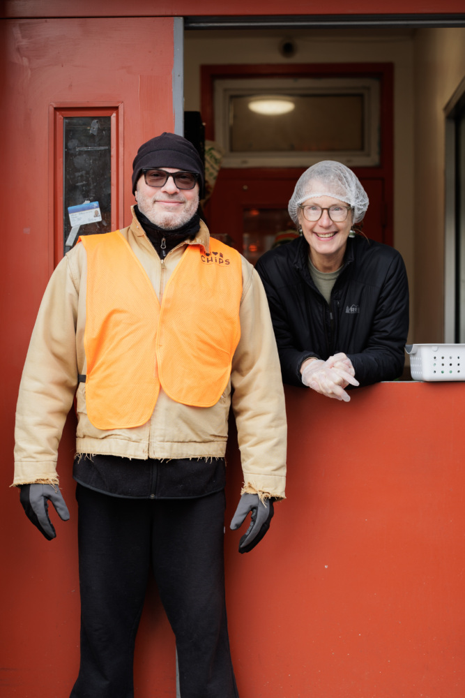 Two people stand at a window. One wears an orange vest and black clothes; the other wears a hairnet and gloves, smiling. They're in a red-painted building.