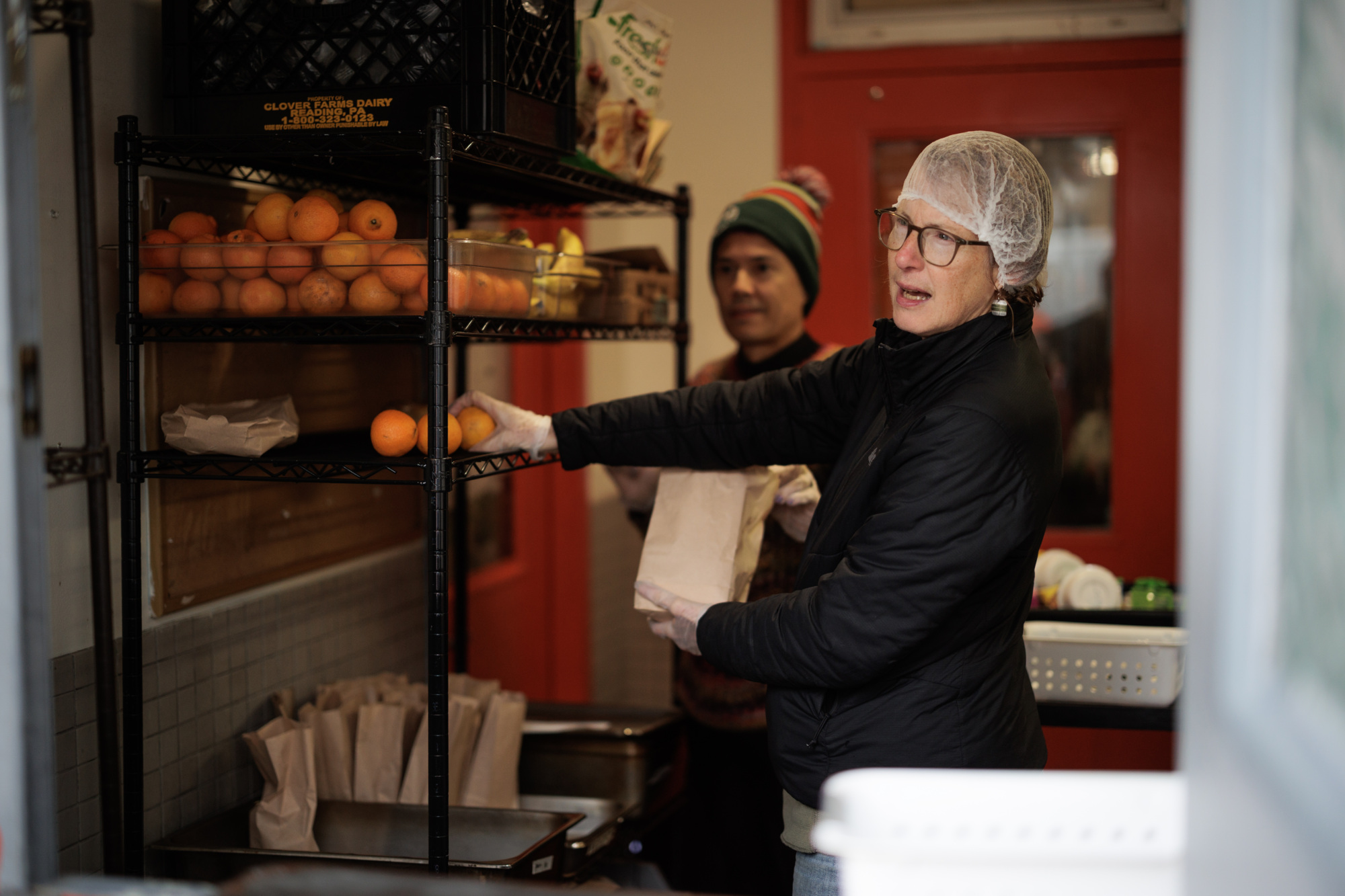 A woman wearing a hairnet and gloves packages food into paper bags from a shelf with oranges. A man stands behind her in a knit cap.