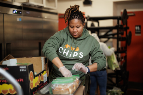 Person in a green hoodie prepares food packages in a kitchen setting.