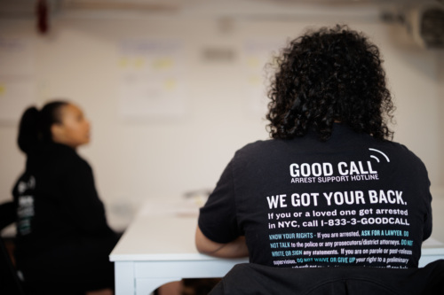 Person wearing a "Good Call Arrest Support Hotline" shirt sits at a table in a room with another person. The shirt provides information and a hotline number for legal assistance in NYC.