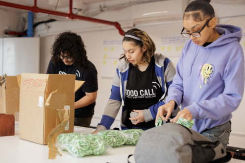 Three people organize phone cords at a table. One person is packing a box labeled "GOOD CALL CORDS," while the others assist. A wall with sticky notes is visible in the background.