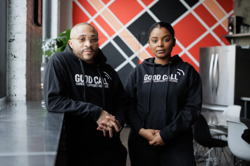 Two people wearing "Good Call Arrest Support Hotline" hoodies stand in a modern room with a geometric red and black wall design.