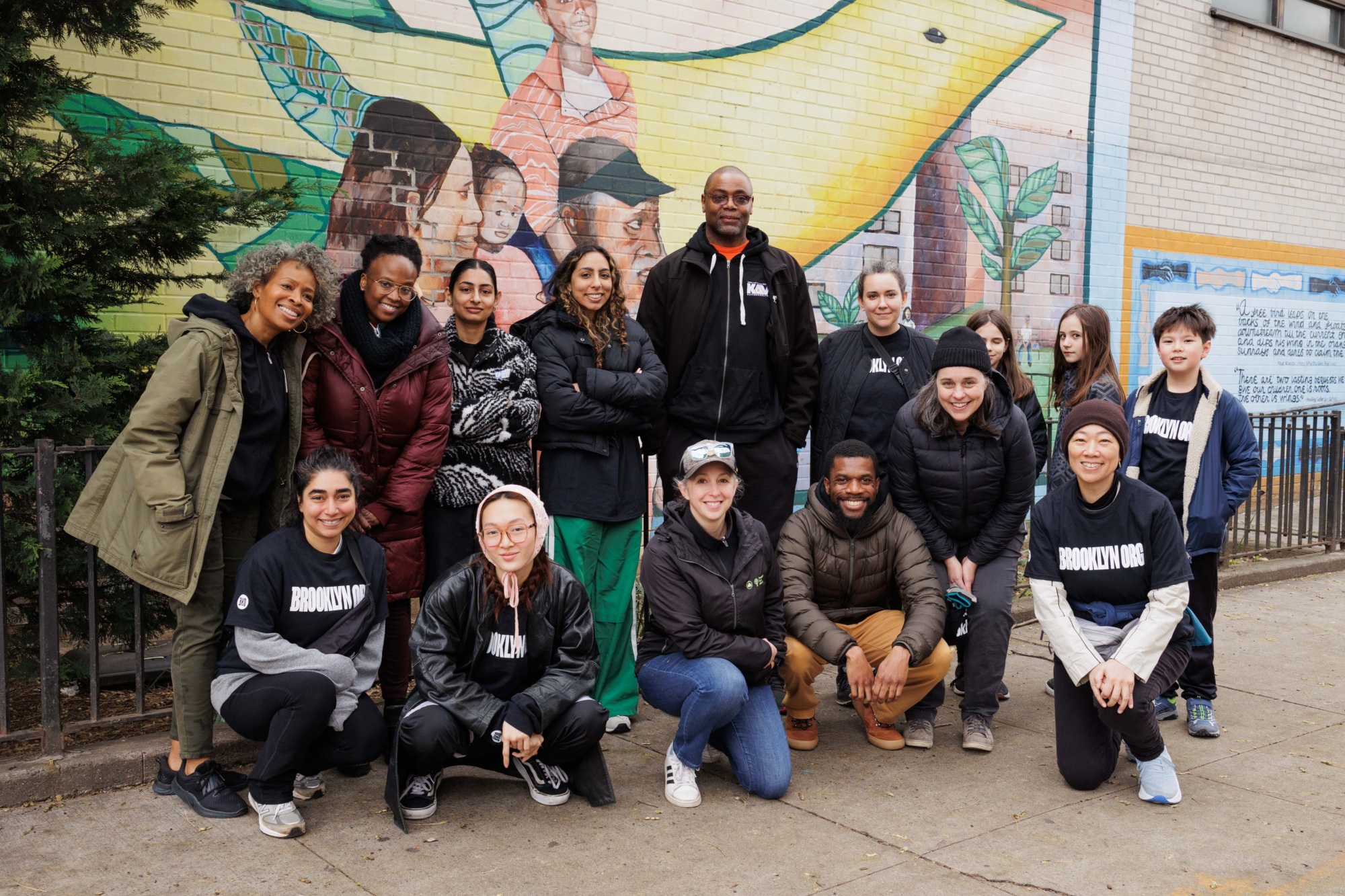 A diverse group of people poses together in front of a colorful mural on an outdoor wall.