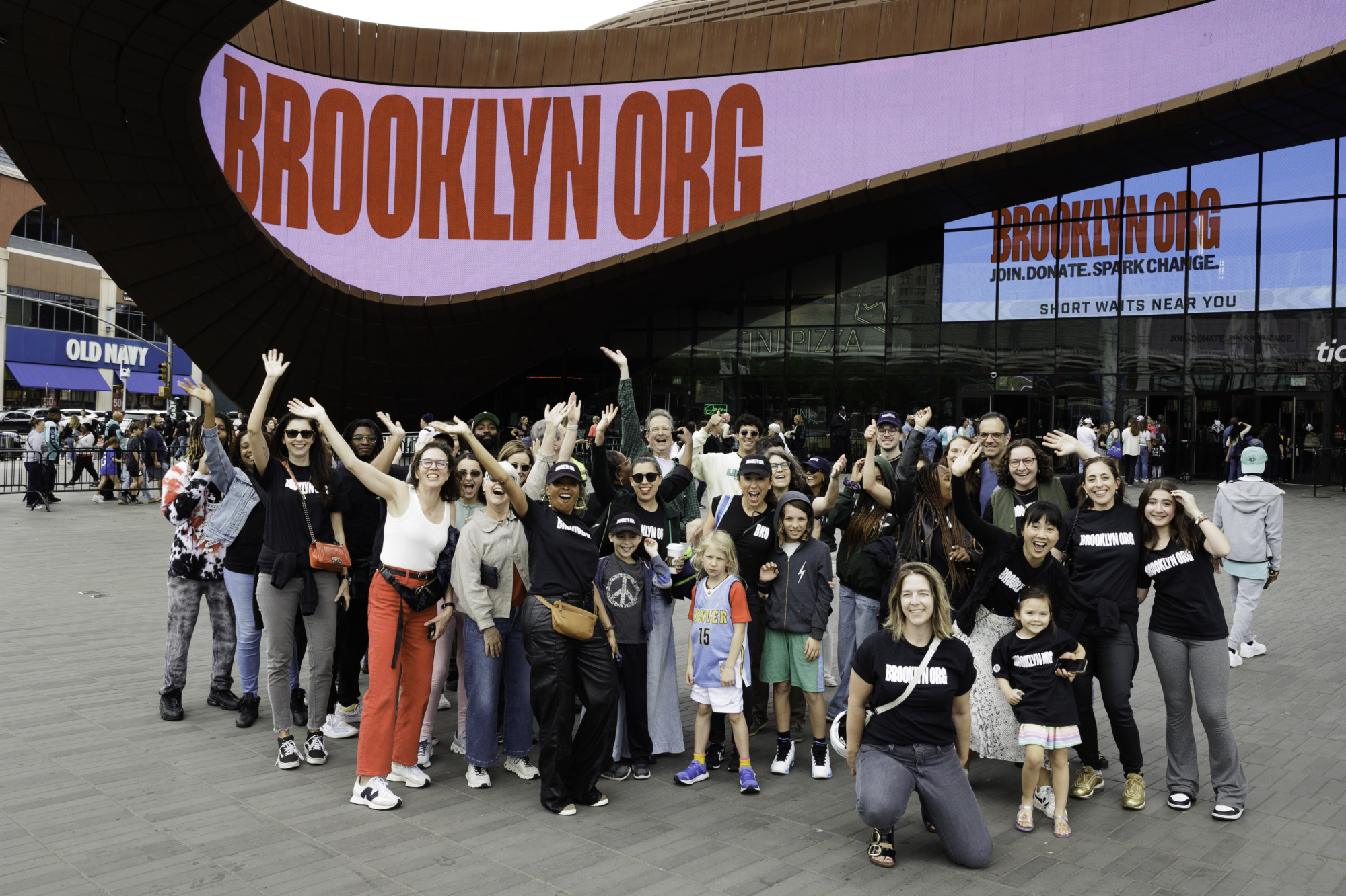 A diverse group of people poses joyfully in front of the Barclays Center, with "BROOKLYN.ORG" displayed on the building behind them.