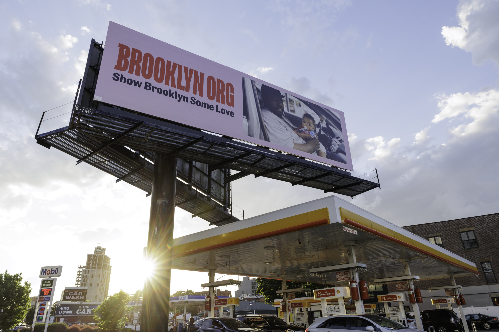 Billboard over a gas station reads "BROOKLYN.ORG Show Brooklyn Some Love," with an image of a person holding a baby.