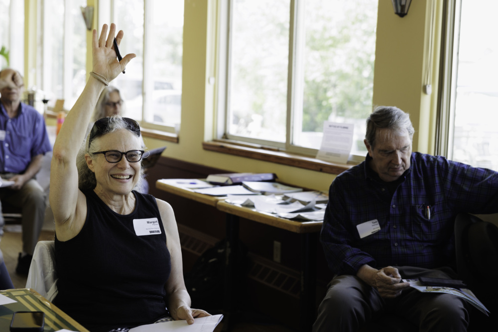 A woman with glasses raises her hand while seated in a room with others. A man sits beside her looking at a notebook.