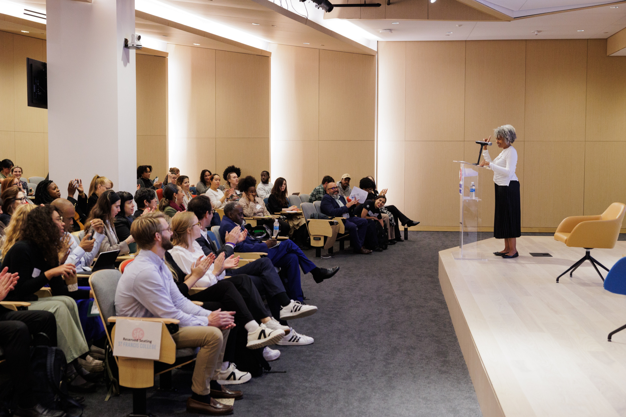 A speaker addresses a seated audience in a conference room. Attendees listen attentively, some taking notes and others applauding.