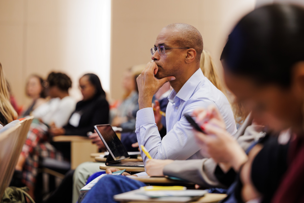 A man sits in a lecture hall, listening attentively, surrounded by other attendees. Some are using electronic devices.