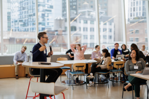 A person speaks into a microphone in front of an audience seated in a modern conference room with large windows.